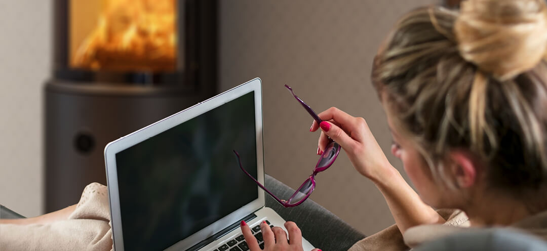 Una mujer con pelo se sienta frente a una estufa de leña Scan 67 negra con una PC en el regazo y gafas en la mano